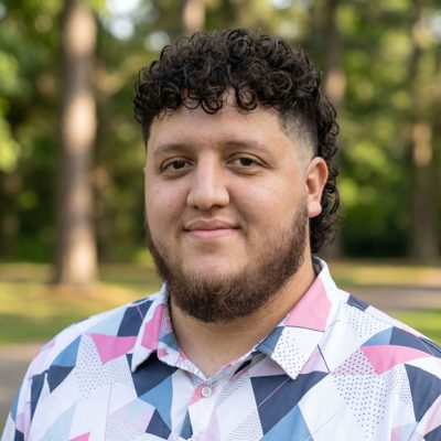 Junior Fortunato — a man with dark curly hair and a short beard, in a pink-and-blue patterned shirt, smiling slightly. Photographed outdoors with trees behind him.