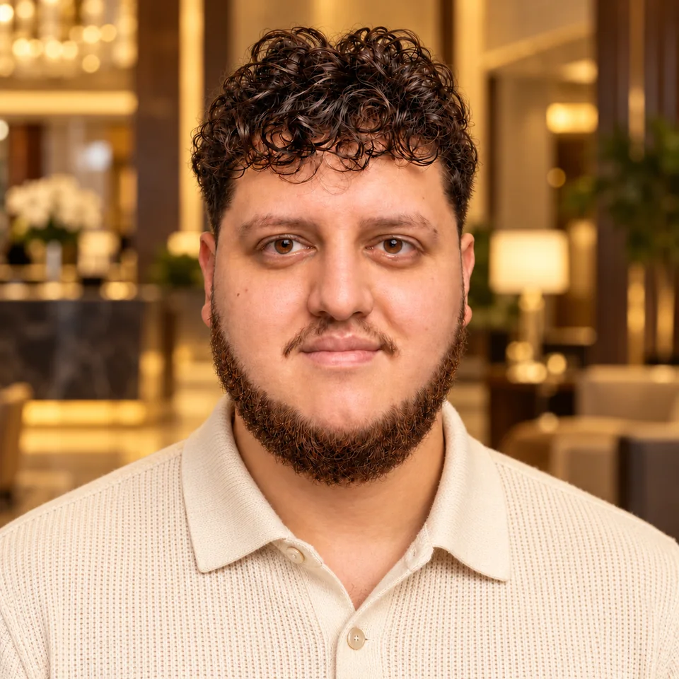 Junior Fortunato with dark curly hair and a beard, wearing a cream knit shirt in a warmly lit lounge.